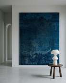 Large abstract blue painting on a white wall with a small wooden stool and white sculpture in front blue rug tapijt designed by wendy guns from ashtari carpets